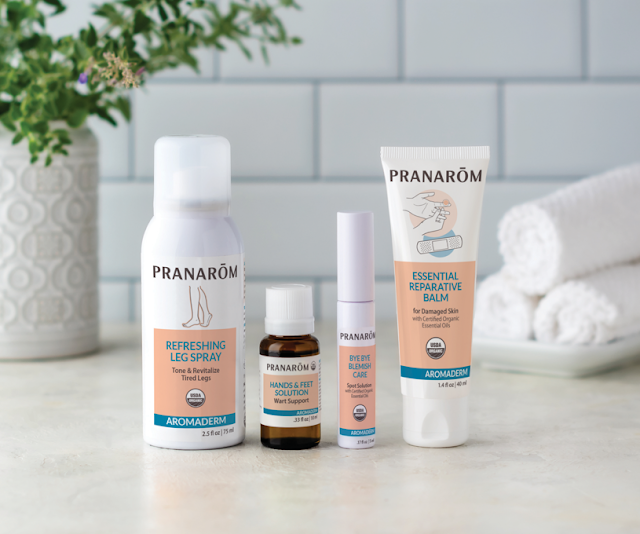 Pranarom Certified Organic Refreshing Leg Spray, Hands & Feet Solution, Bye Bye Blemish Care, and Essential Reparative Balm Aromaderm bottles with towels and green plant on white background.