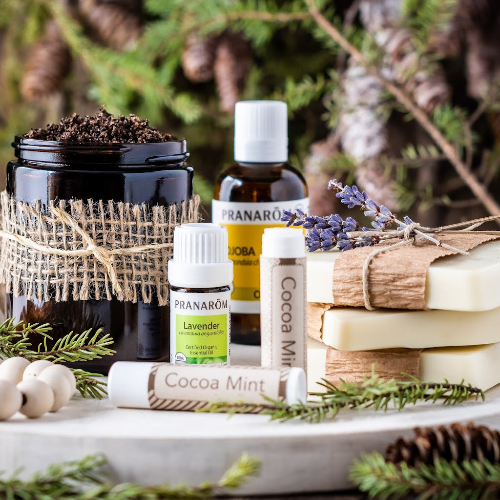 Pranarom Certified Organic Lavender Essential Oil, Cocoa Mint Lip Balm, and Jojoba Virgin Plant Oil bottles with nature background and bar soaps.